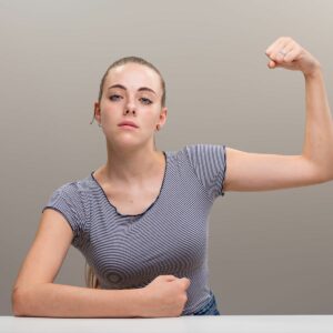 A confident woman in a striped shirt flexes her arm, showing strength, while sitting at a table against a neutral background.