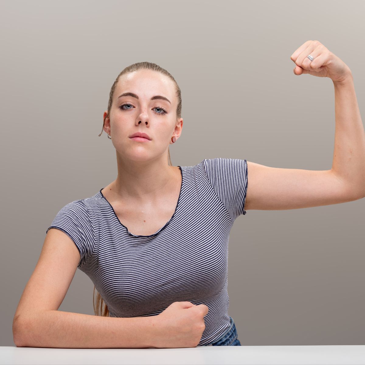 A confident woman in a striped shirt flexes her arm, showing strength, while sitting at a table against a neutral background.