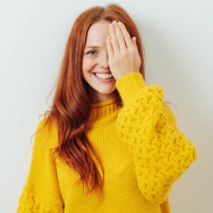 A smiling woman with long red hair wearing a yellow sweater covers one eye with her hand while standing against a white background.