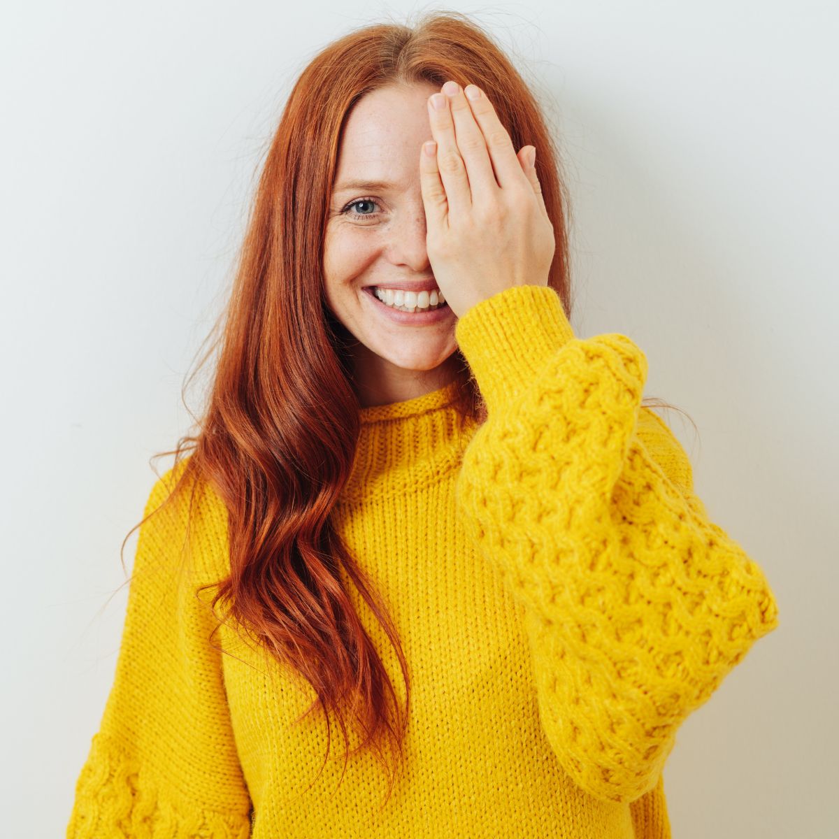 A smiling woman with long red hair wearing a yellow sweater covers one eye with her hand while standing against a white background.