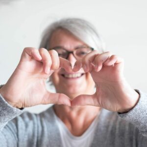 An older woman with short gray hair and glasses making a heart shape with her hands, smiling in the background, wearing a gray sweater.