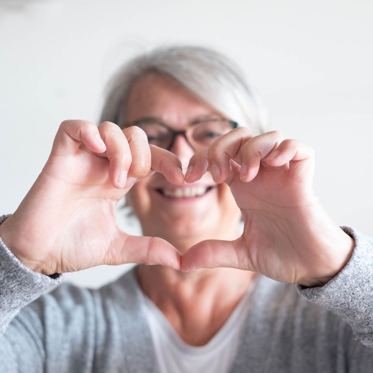 An older woman with short gray hair and glasses making a heart shape with her hands, smiling in the background, wearing a gray sweater.
