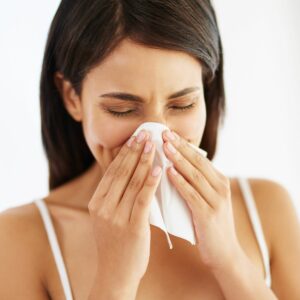 A woman sneezing into a tissue, with her eyes closed and a pained expression, suggesting she may have a cold or allergies.