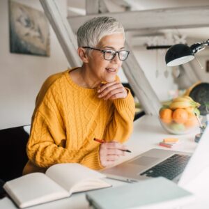 An older woman with short gray hair and glasses wearing a yellow sweater sits at a desk, smiling and working on a laptop with a notebook and a bowl of fruit nearby.