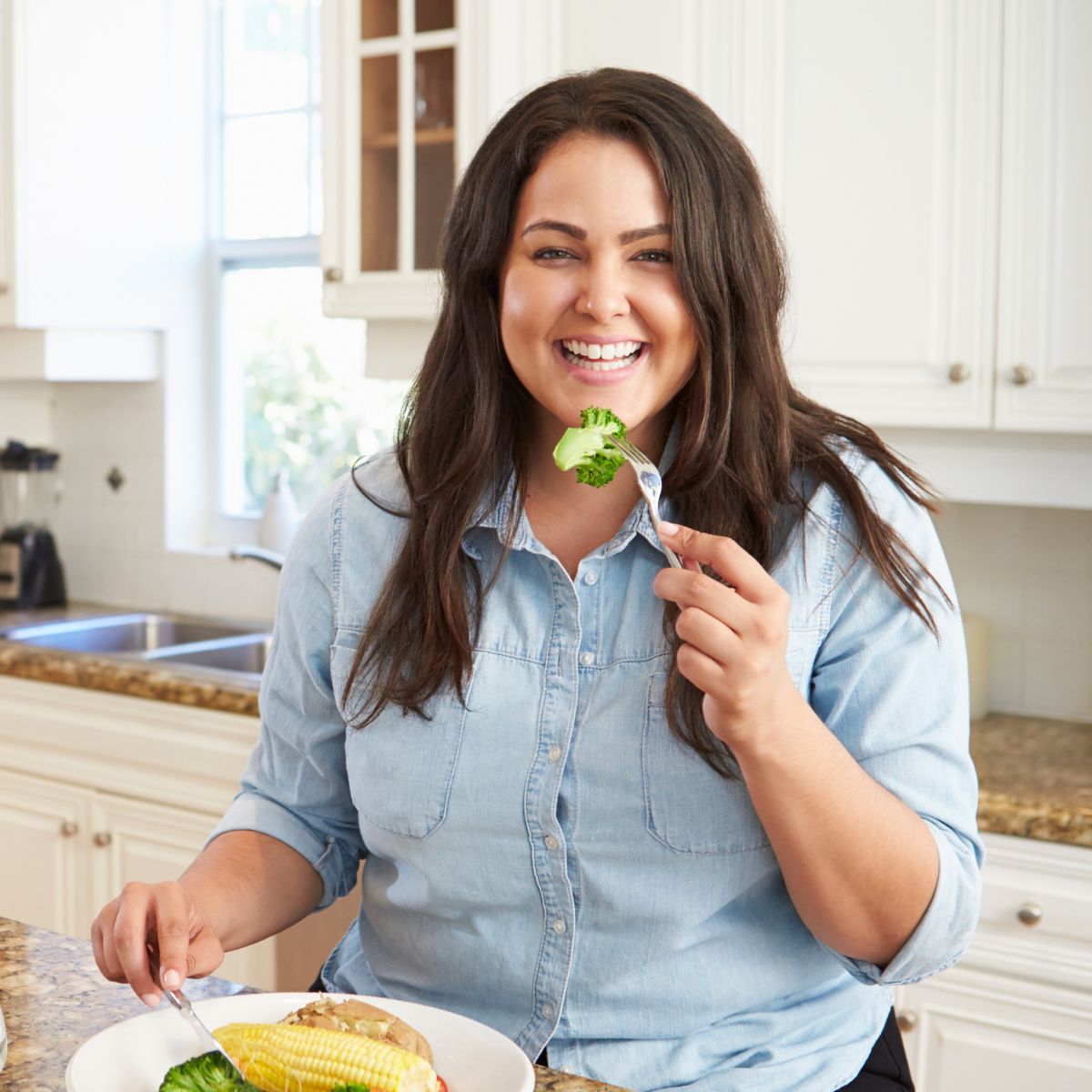 A smiling woman in a denim shirt enjoys a healthy meal in her kitchen, holding a fork with a piece of broccoli.